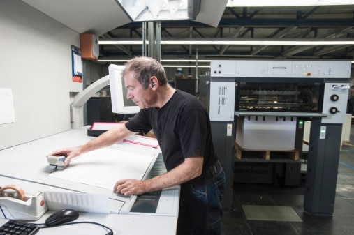 Press operator checking freshly printed sheet from a sheetfed offset press in a viewing booth with color-controlled lighting.